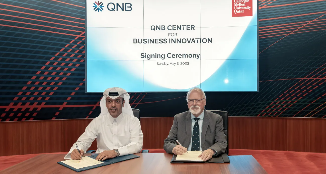 Two men seated at a conference table sign documents during a signing ceremony, one in a white thobe and the other in a suit, with a large screen behind them reading 'QNB Center for Business Innovation Signing Ceremony'