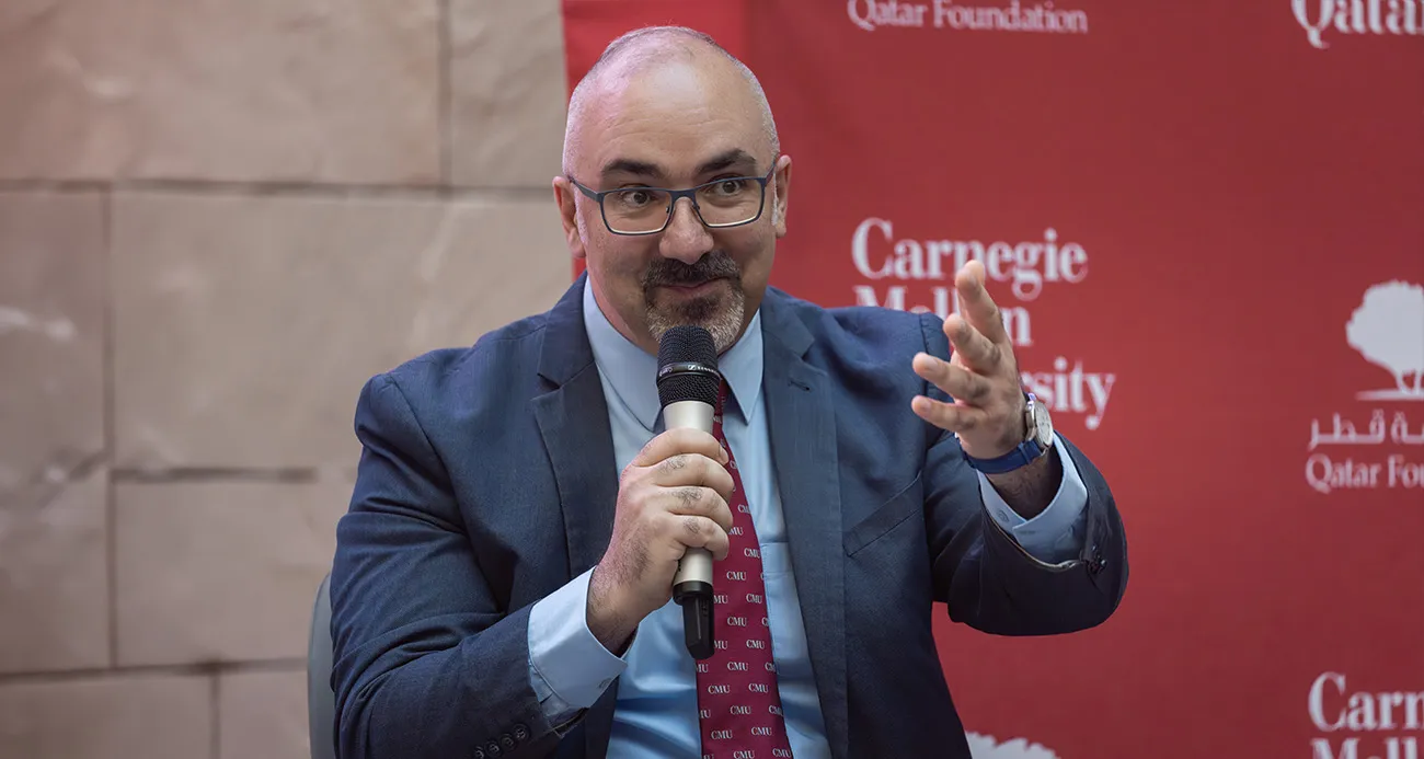 Man in a suit and glasses speaks into a handheld microphone, gesturing with his left hand on a red backdrop.