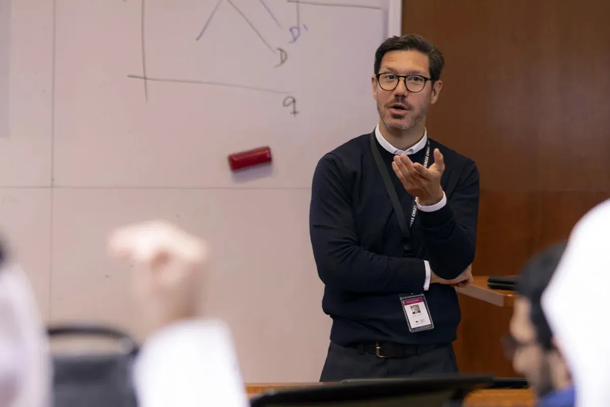 Educator in a dark sweater with a lanyard teaches at the front, gesturing as students watch, with a scribbled whiteboard behind him.